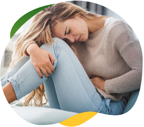 A young woman suffering from period bloating and sitting cramped on the floor holding her abdomen with her left hand